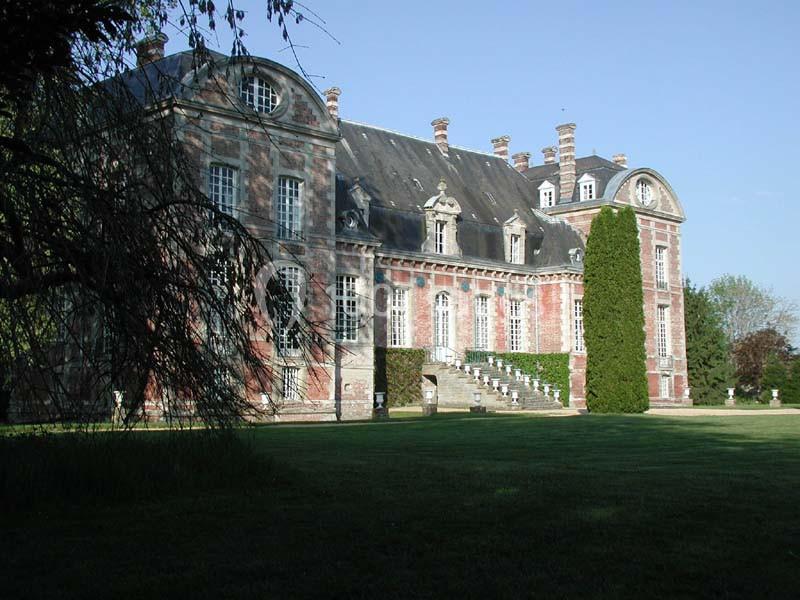 Façade d'un château en briques rouges avec toit en ardoise, entouré de pelouse et d'arbres sous un ciel dégagé.