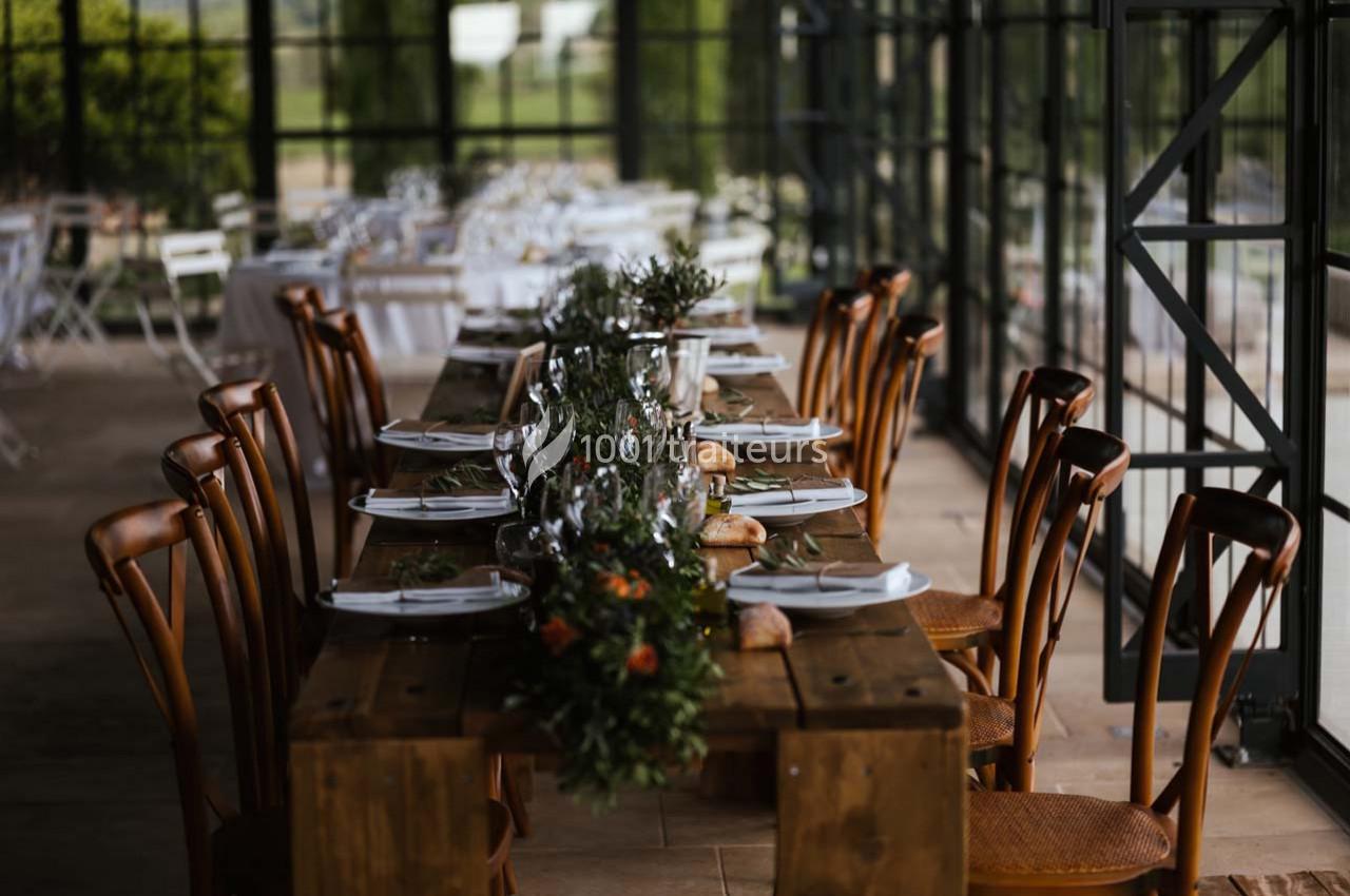 Table en bois dressée avec vaisselle, verres et décoration florale, entourée de chaises dans une salle vitrée.