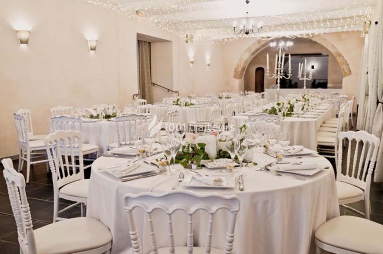 Salle de réception décorée pour un mariage, avec tables rondes dressées, chaises blanches et guirlandes lumineuses.