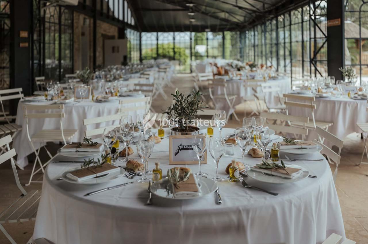 Salle lumineuse avec tables rondes dressées pour un repas, décorées de nappes blanches et centres de table végétaux.