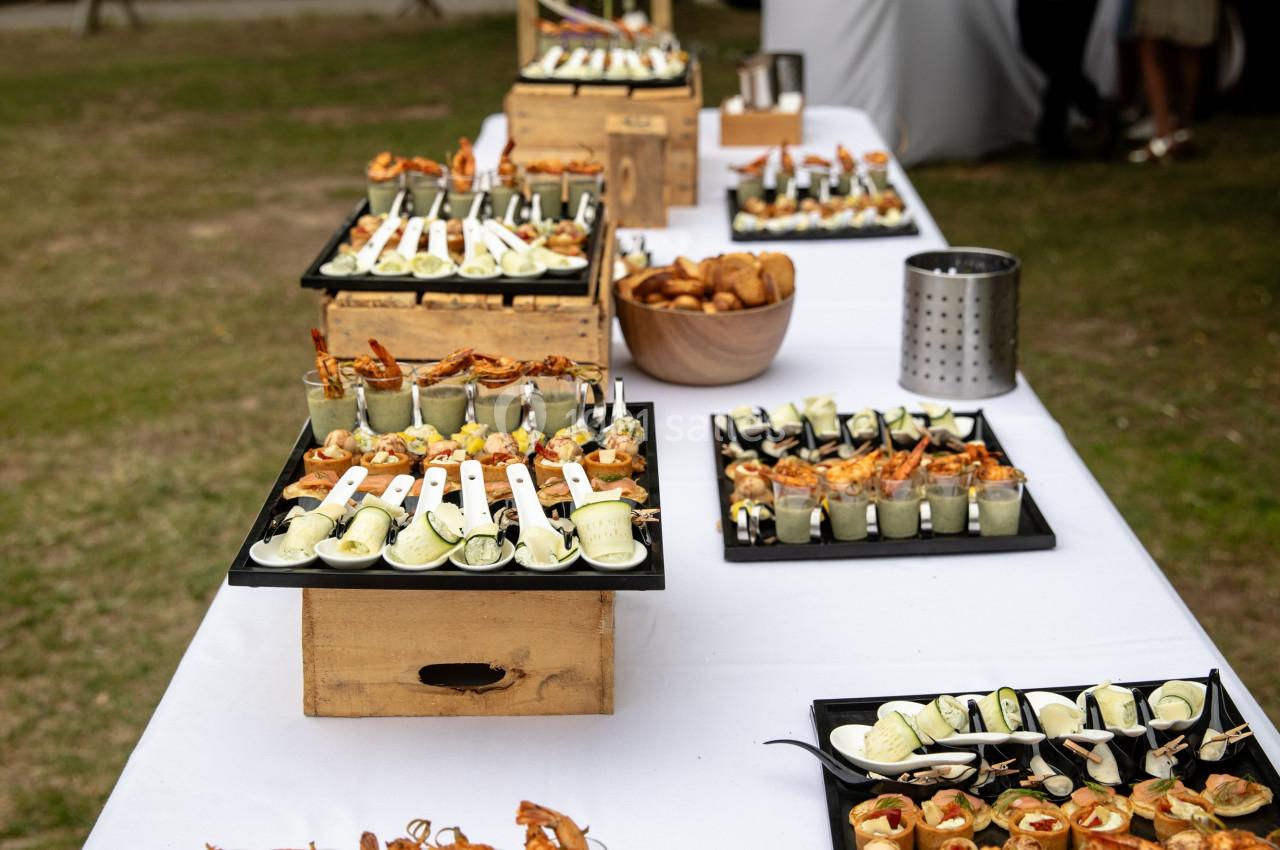 Buffet en plein air avec verrines, cuillères apéritives et amuse-bouches disposés sur une longue table blanche.