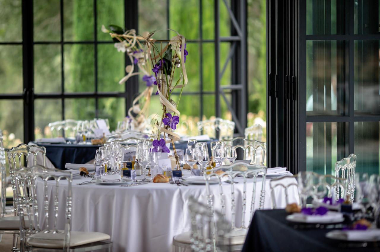 Salle de réception avec tables élégamment dressées, décorées de fleurs violettes et chaises transparentes.