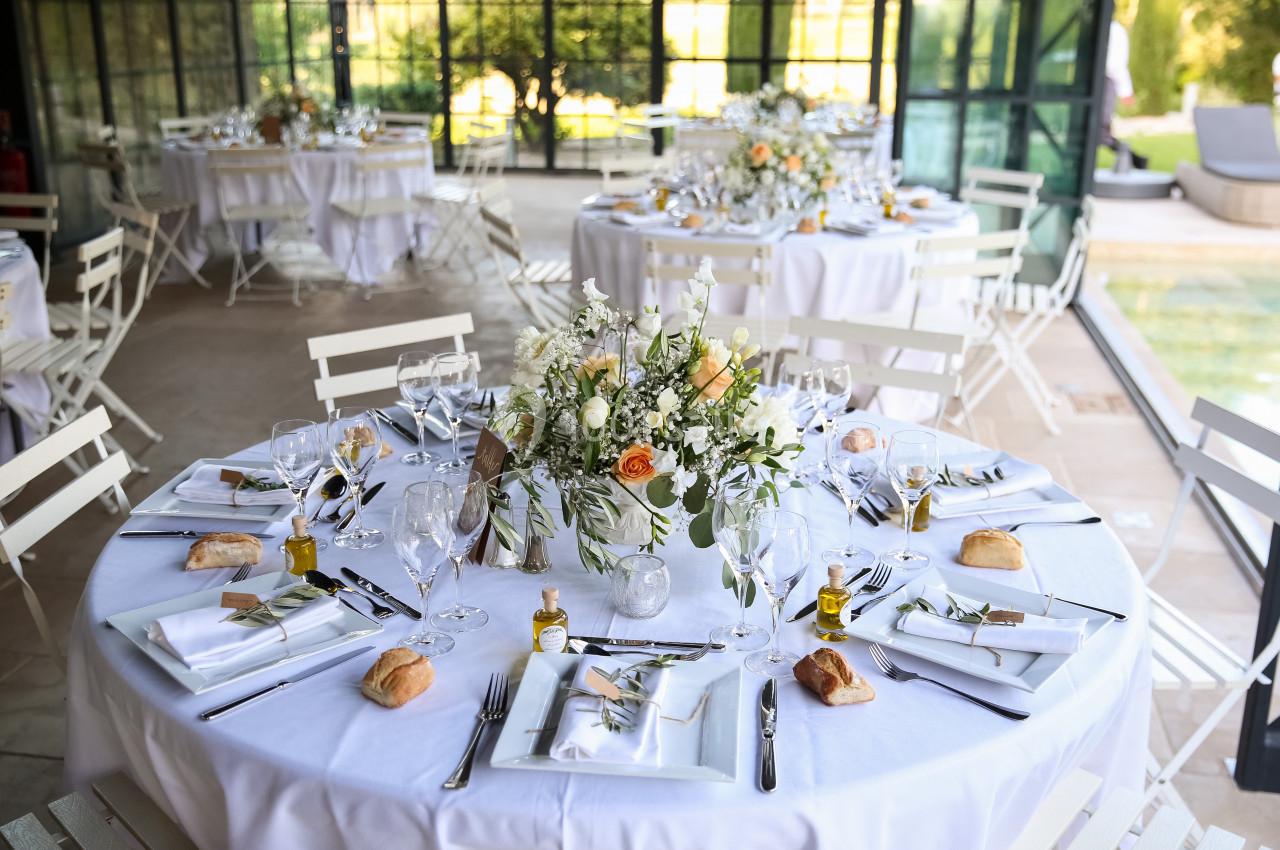 Tables rondes élégamment dressées avec nappes blanches, bouquets floraux et couverts, dans une salle lumineuse avec vue sur…