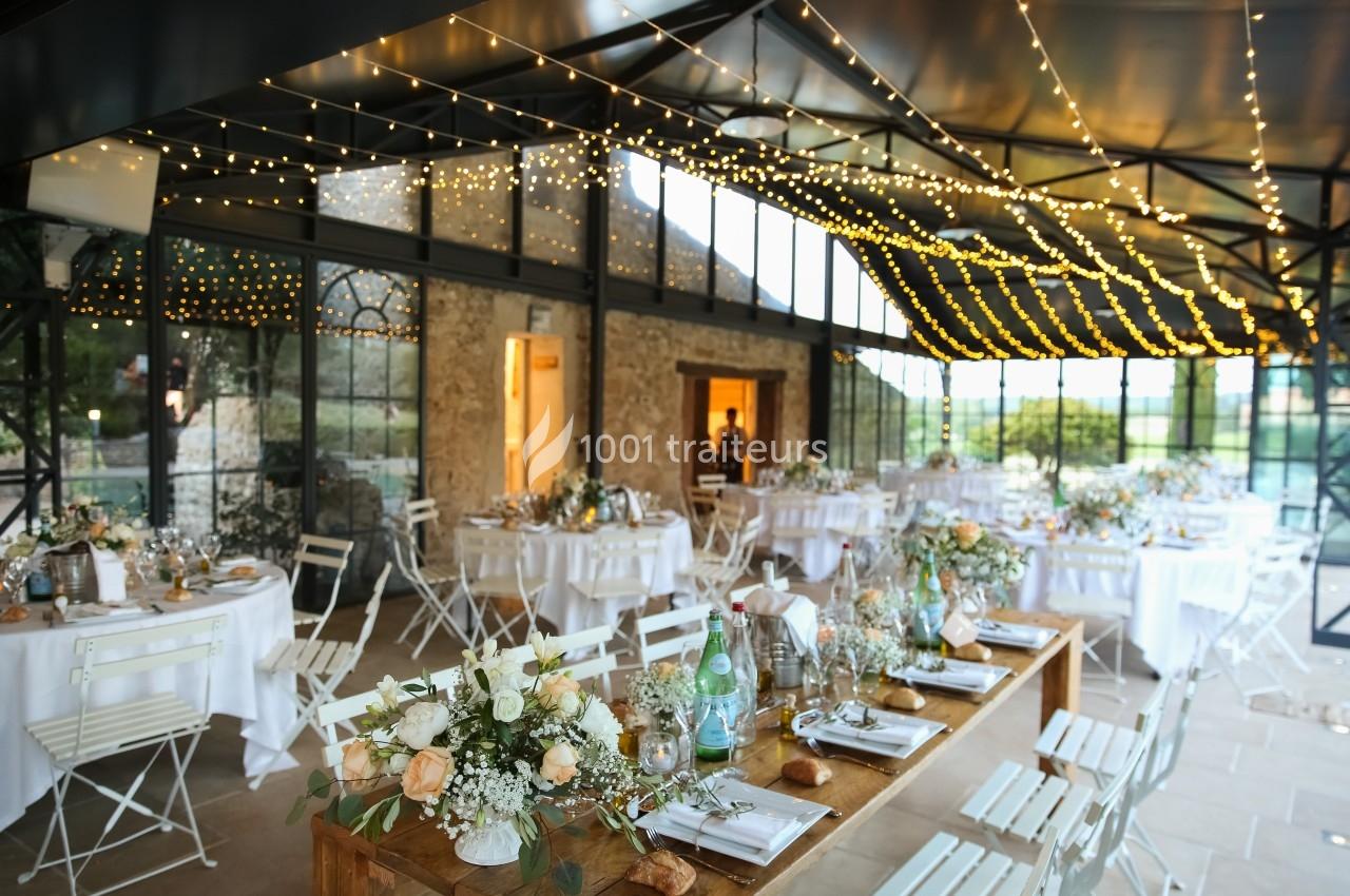 Salle de réception décorée pour un mariage, avec tables dressées, guirlandes lumineuses et grandes baies vitrées.
