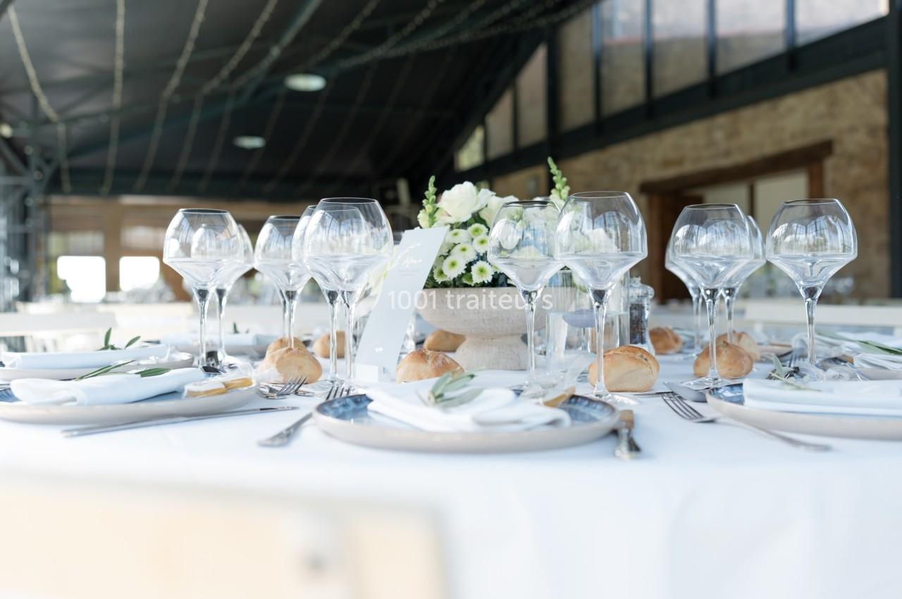 Table dressée avec nappes blanches, verres à vin, couverts, petits pains et centre de table floral dans une salle lumineuse.