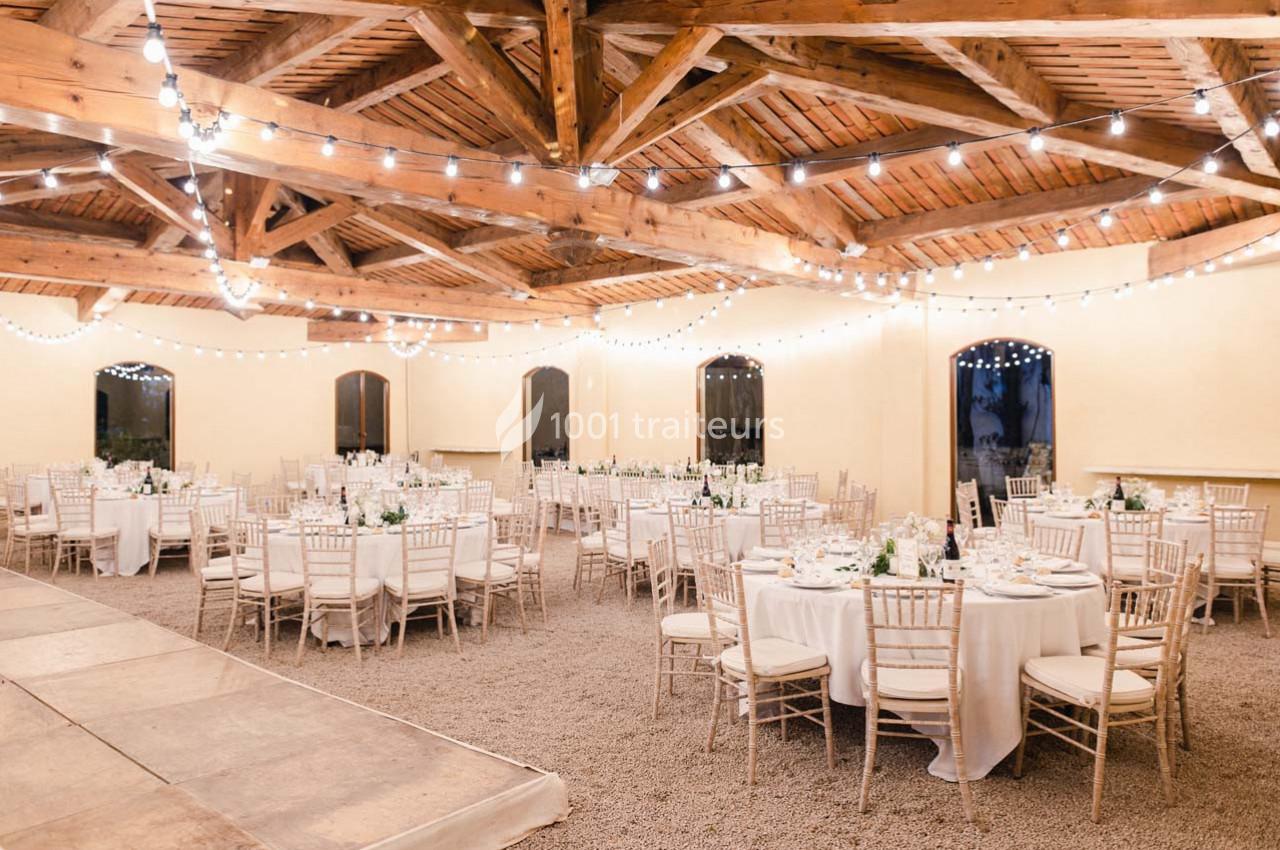Salle de réception lumineuse avec tables rondes dressées, chaises en bois clair et guirlandes suspendues au plafond en bois.