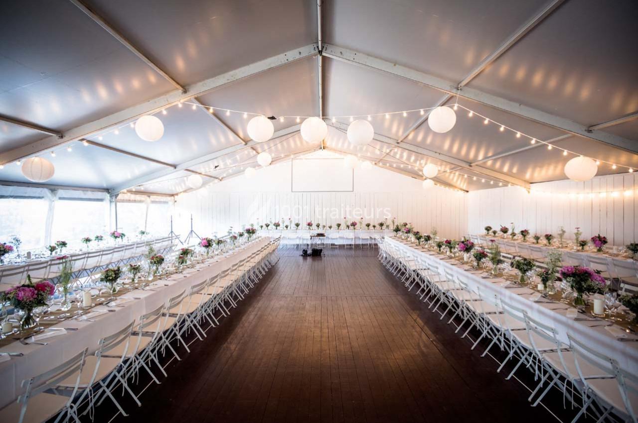 Grande salle de réception sous une tente, avec de longues tables décorées de fleurs et éclairées par des guirlandes…