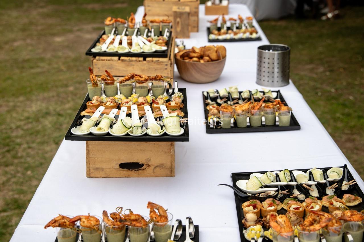 Buffet en extérieur avec des plateaux de verrines, brochettes et amuse-bouches variés sur une nappe blanche.