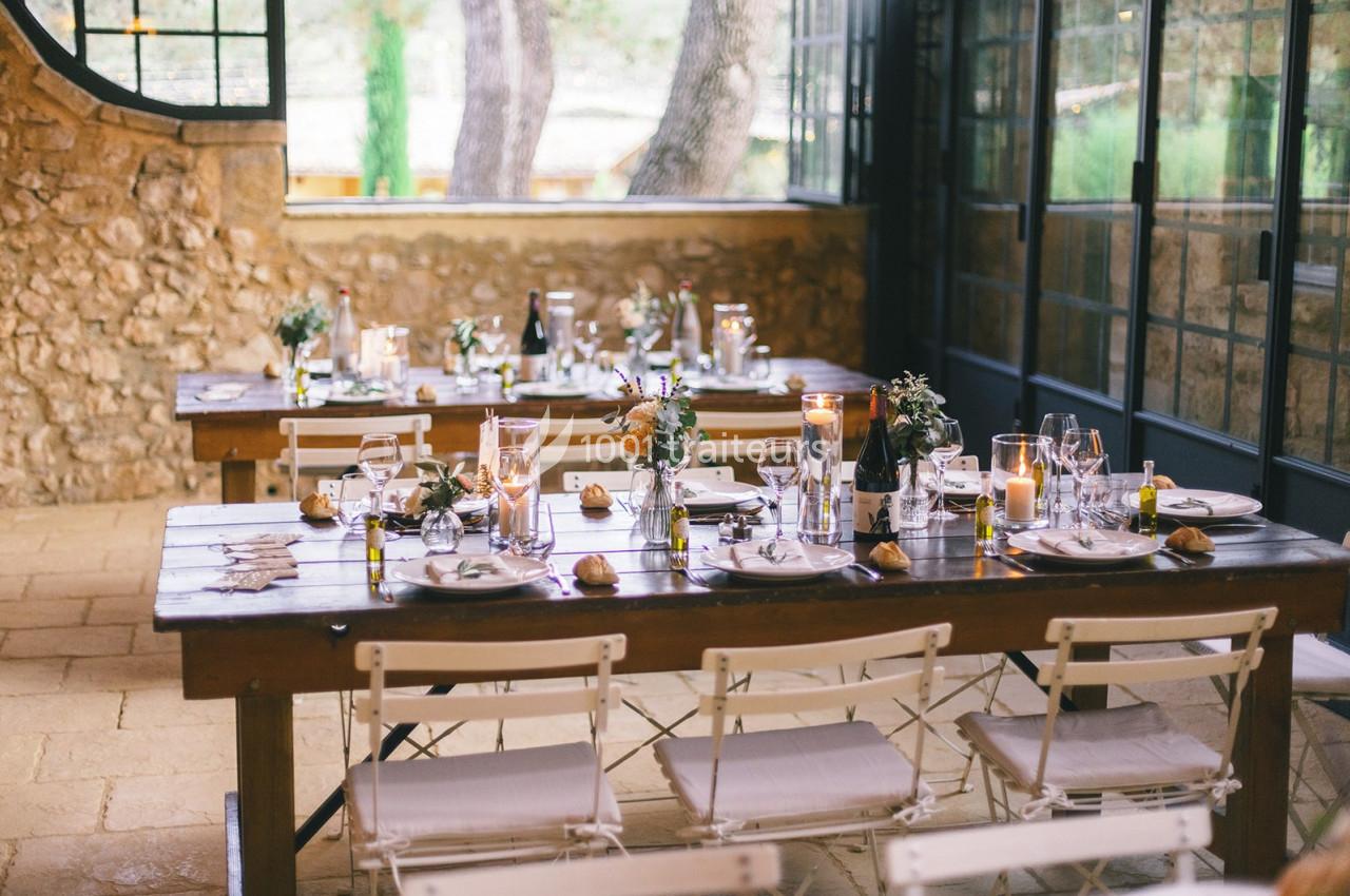 Tables en bois dressées avec vaisselle, bougies et décorations florales dans une salle lumineuse en pierre.