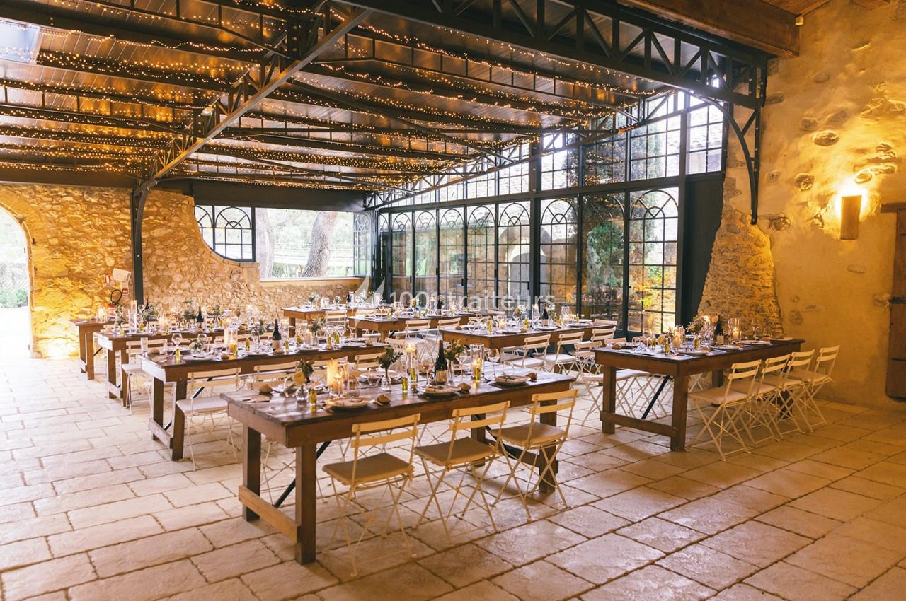 Salle de réception lumineuse avec tables en bois, chaises blanches et guirlandes suspendues sous un toit en verrière.
