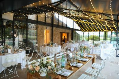 Une mariée en robe blanche tient un bouquet dans une salle décorée pour un mariage, avec un homme en costume bleu derrière…