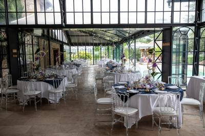 Une mariée en robe blanche tient un bouquet dans une salle décorée pour un mariage, avec un homme en costume bleu derrière…