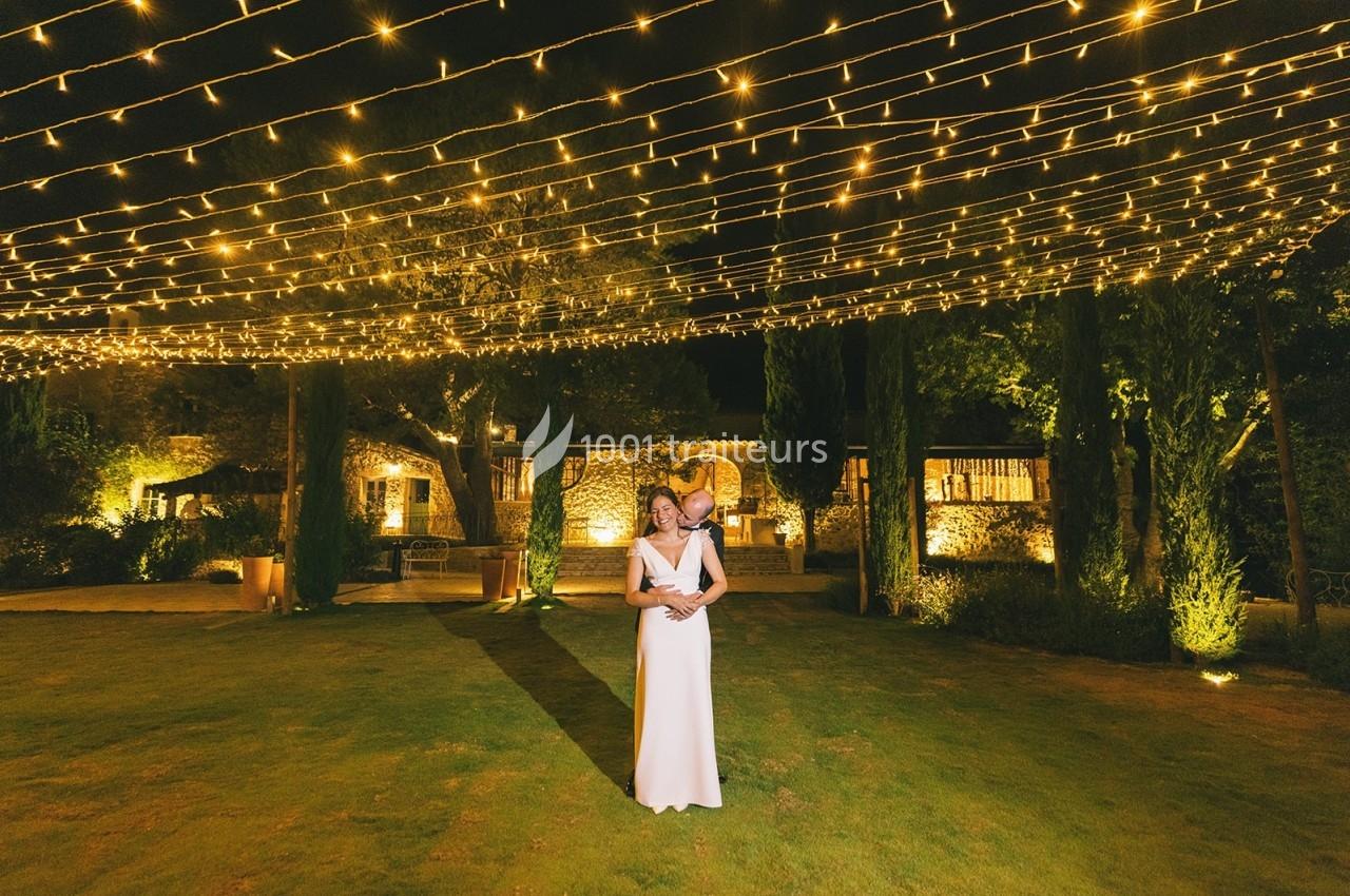 Un couple pose sous des guirlandes lumineuses dans un jardin éclairé, entouré de végétation et de bâtiments en pierre.