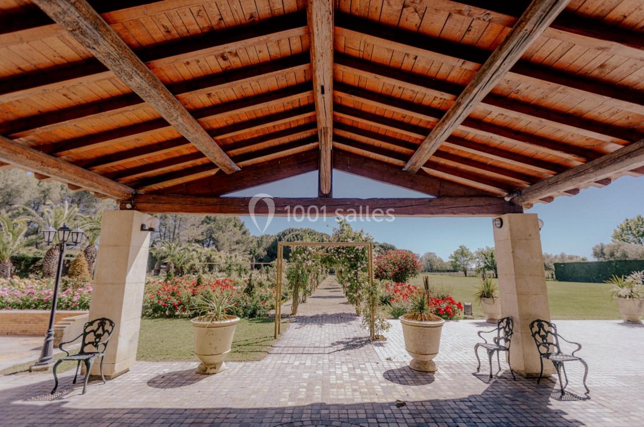 Location salle Marsillargues (Hérault) - Le Mas Saint Gabriel #6 Structure en bois avec toit ouvert, entourée de jardins fleuris et d'une allée centrale bordée de verdure.