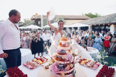Miniature Location salle Marsillargues (Hérault) - Le Mas Saint Gabriel #13 Une mariée souriante brandit un éventail devant un gâteau de mariage décoré, entourée d'invités en extérieur.