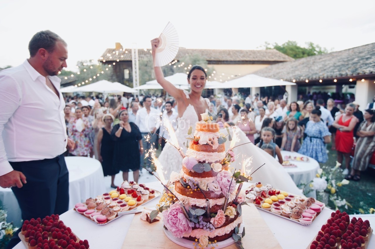Location salle Marsillargues (Hérault) - Le Mas Saint Gabriel #13 Une mariée souriante brandit un éventail devant un gâteau de mariage décoré, entourée d'invités en extérieur.