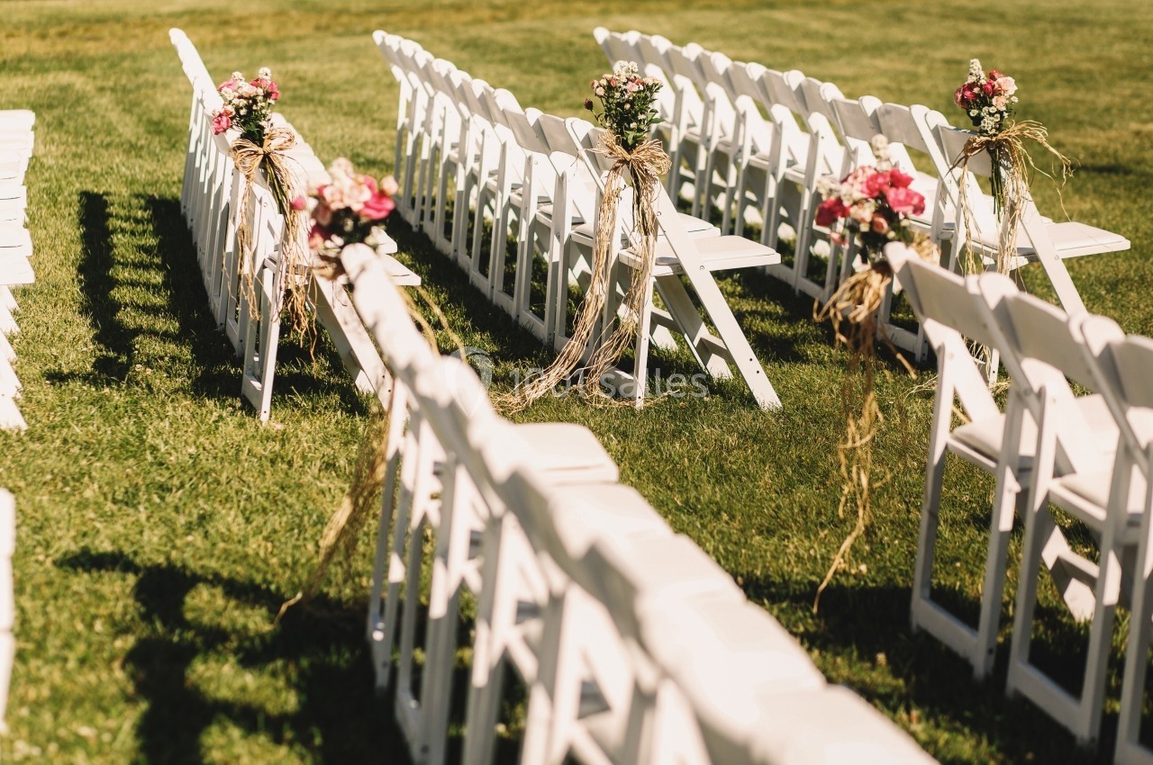 Location salle Marsillargues (Hérault) - Le Mas Saint Gabriel #12 Chaises blanches alignées en extérieur, décorées de bouquets de fleurs roses et de rubans, sur une pelouse verte.