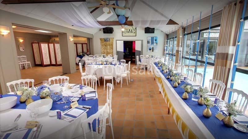 Salle de réception décorée avec des tables dressées, des fleurs bleues et blanches, et une vue sur une piscine.