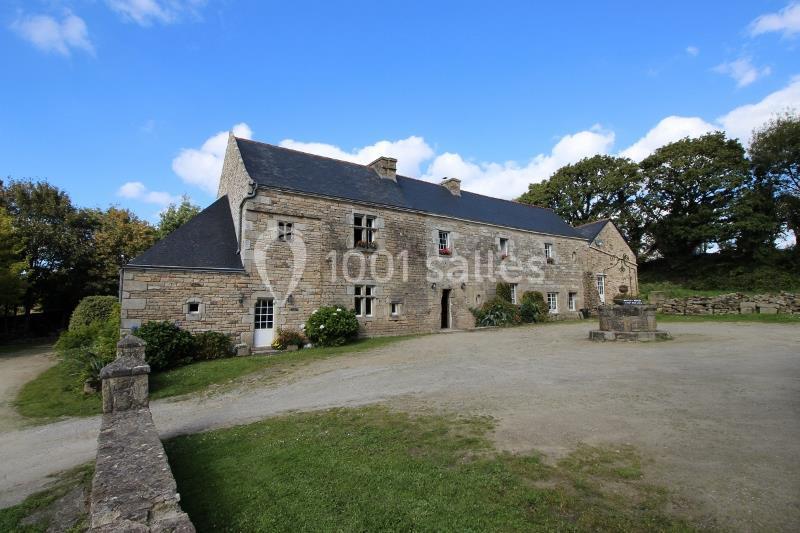 Grande maison en pierre avec toit en ardoise, entourée de végétation et située dans un cadre rural sous un ciel bleu.