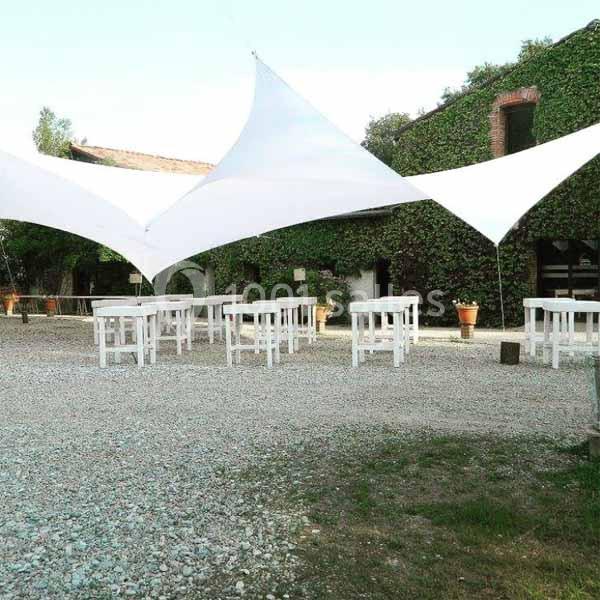 Espace extérieur avec tables et chaises blanches sous des voiles d'ombrage, entouré de bâtiments couverts de végétation.