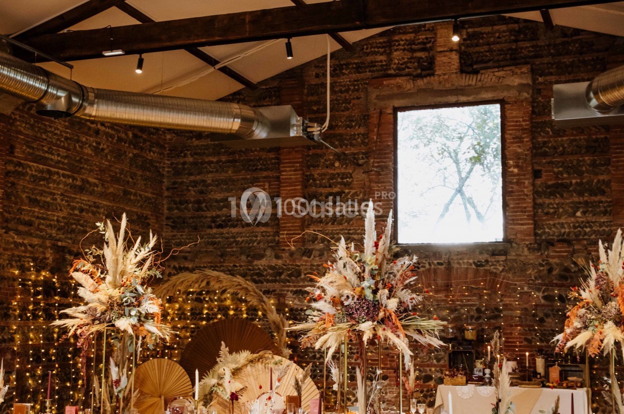 Salle décorée pour un événement, avec des compositions florales séchées, des guirlandes lumineuses et un mur en briques…