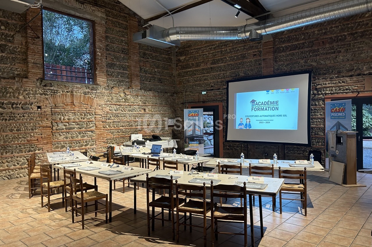 Salle de formation en briques avec tables disposées en U, écran de projection allumé et matériel pédagogique installé.