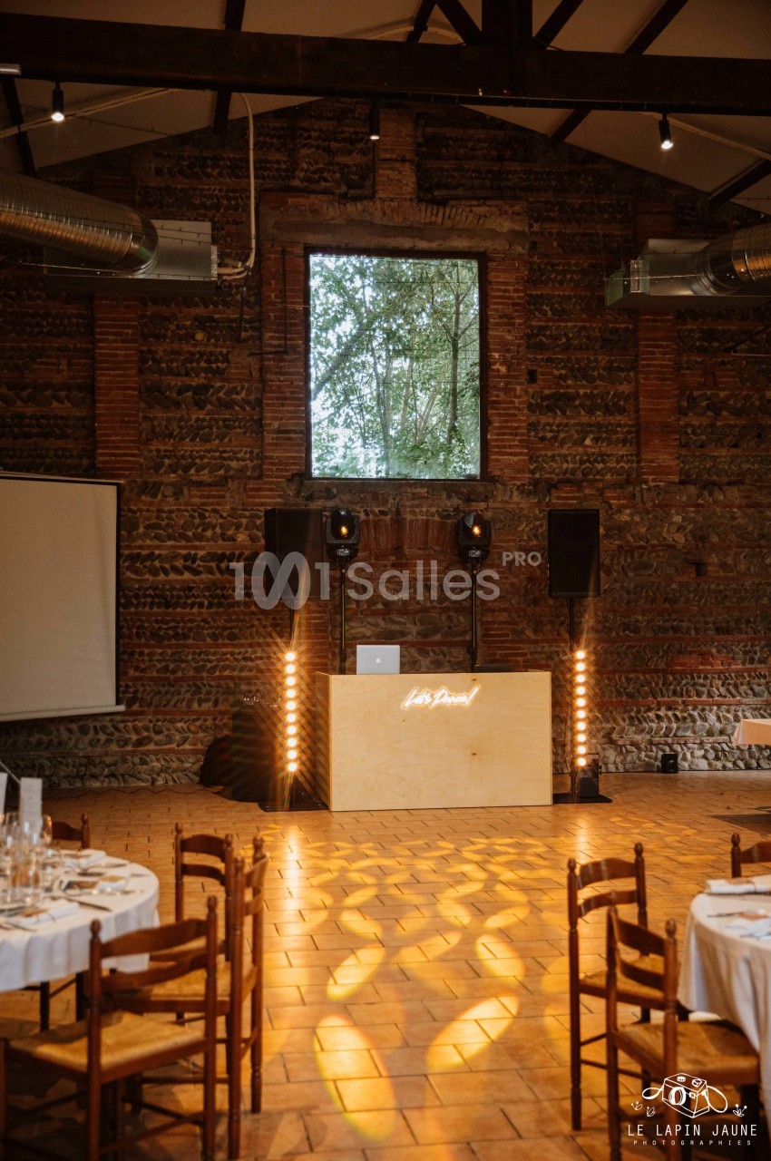 Salle en briques avec une table de DJ éclairée, des chaises en bois et des tables dressées pour un événement.
