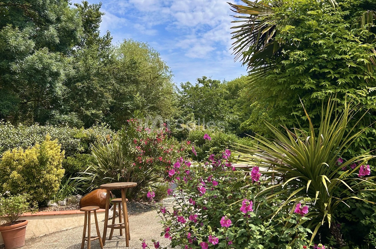 Jardin verdoyant avec des fleurs roses, des arbustes et une petite table en bois sous un ciel partiellement nuageux.