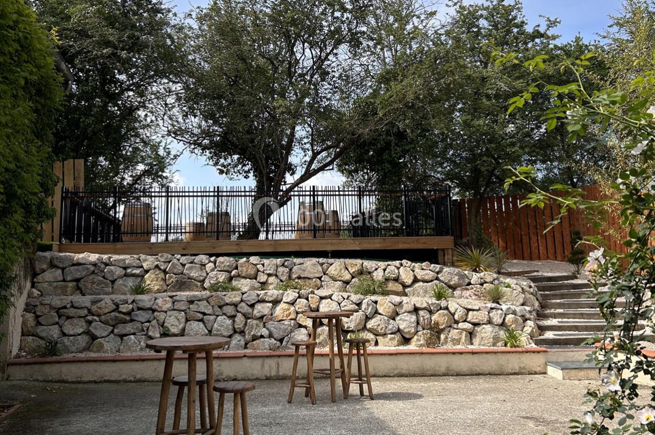 Terrasse surélevée entourée d'arbres, avec des tables en bois sur un sol pavé et un muret en pierres en premier plan.