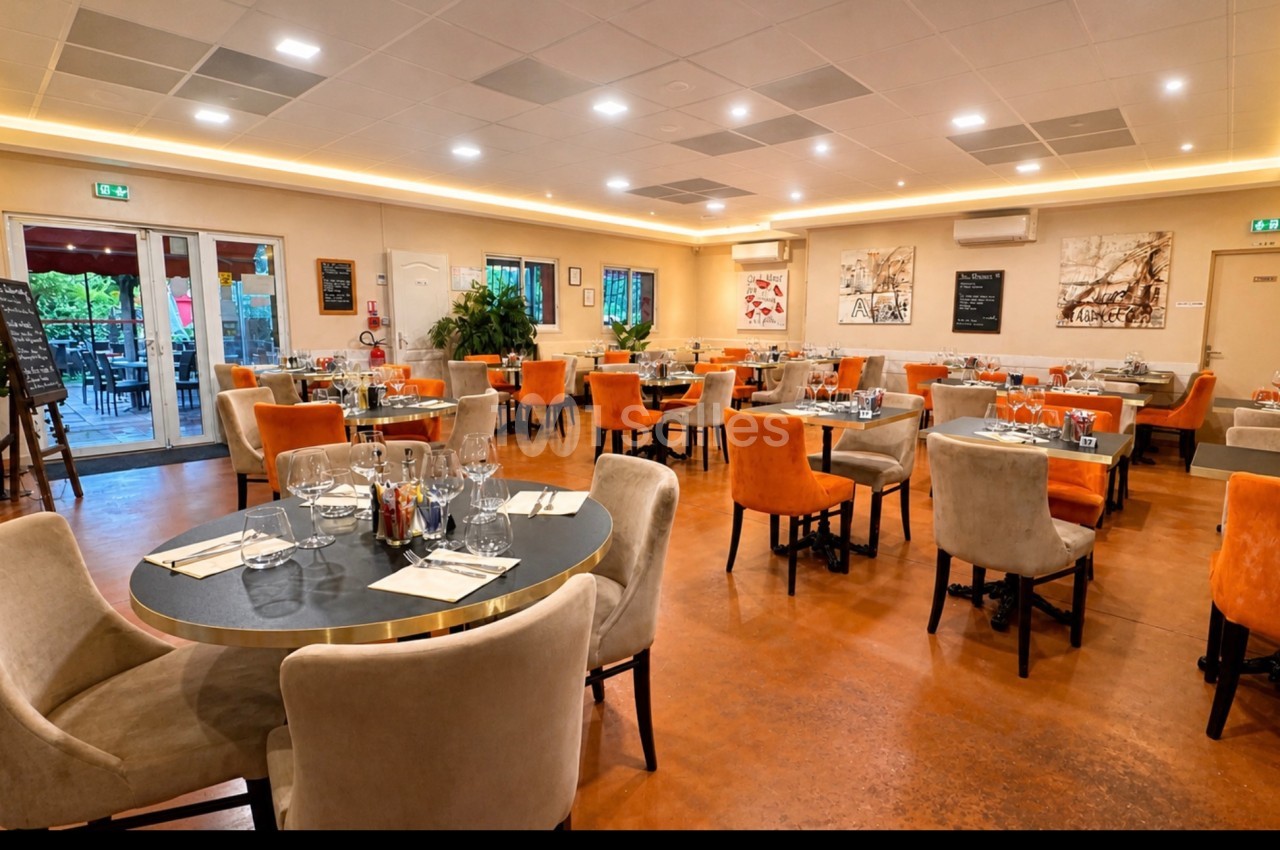 Salle de restaurant lumineuse avec tables dressées, chaises colorées et grandes baies vitrées donnant sur une terrasse.