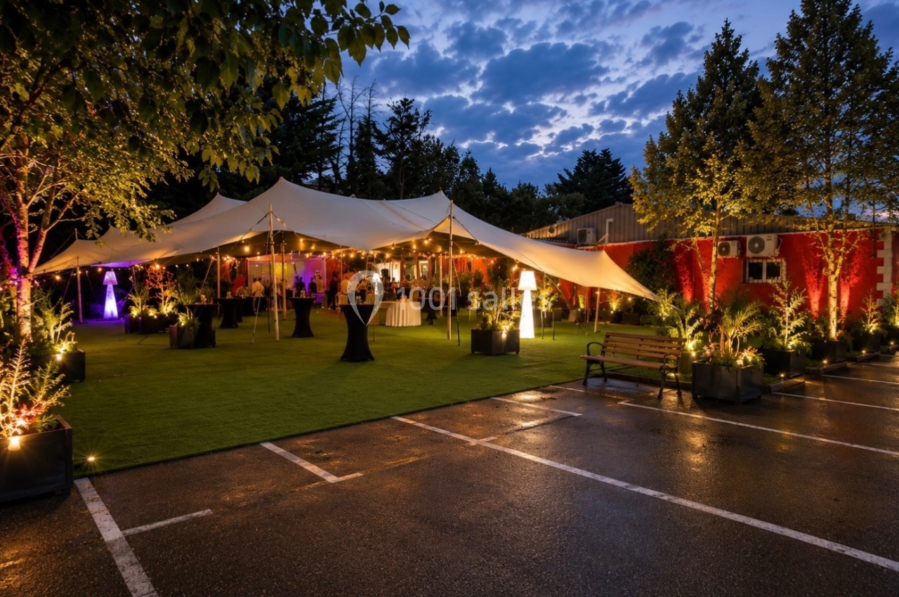 Espace extérieur éclairé avec tente blanche, tables hautes et végétation, dans un cadre de soirée.