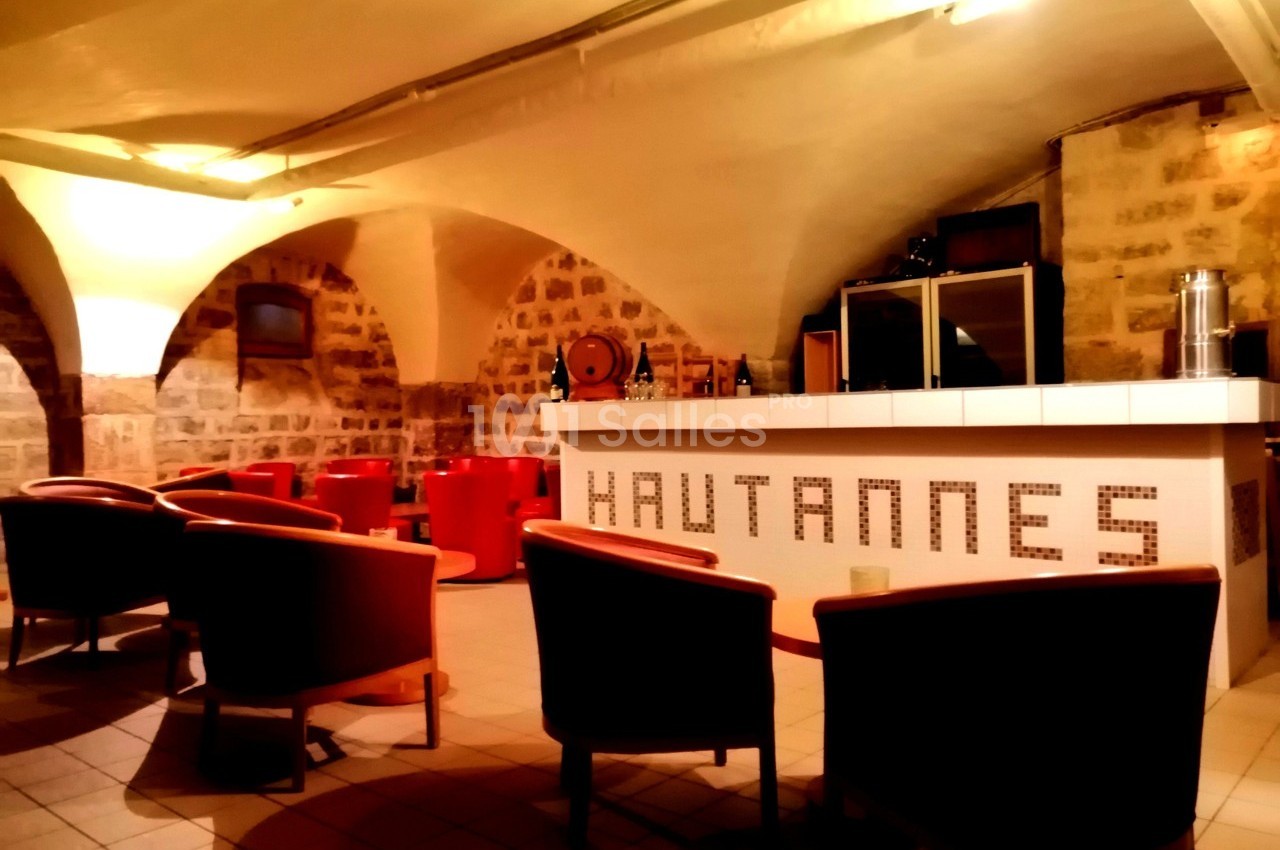 Salle voûtée avec des murs en pierre, un bar blanc marqué ’Hautannes’ et des fauteuils rouges disposés autour de tables.