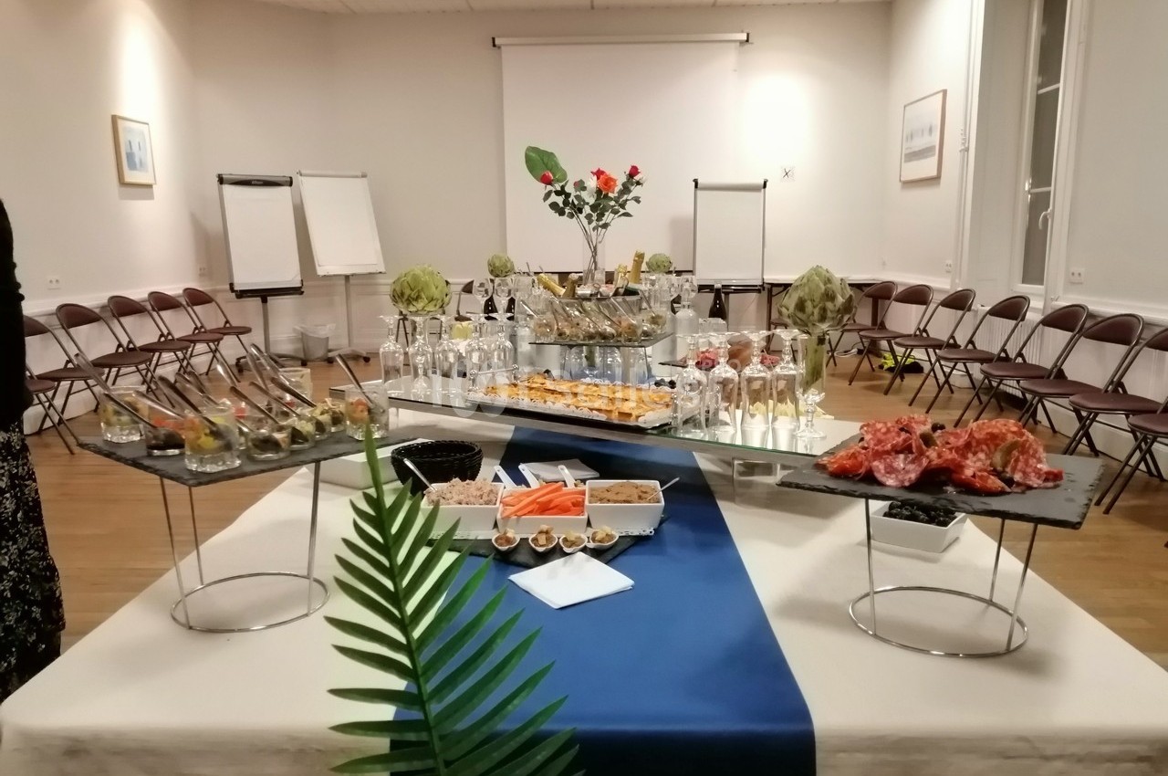 Table dressée pour un buffet avec verrines, charcuterie, légumes, et boissons, dans une salle avec chaises disposées en…