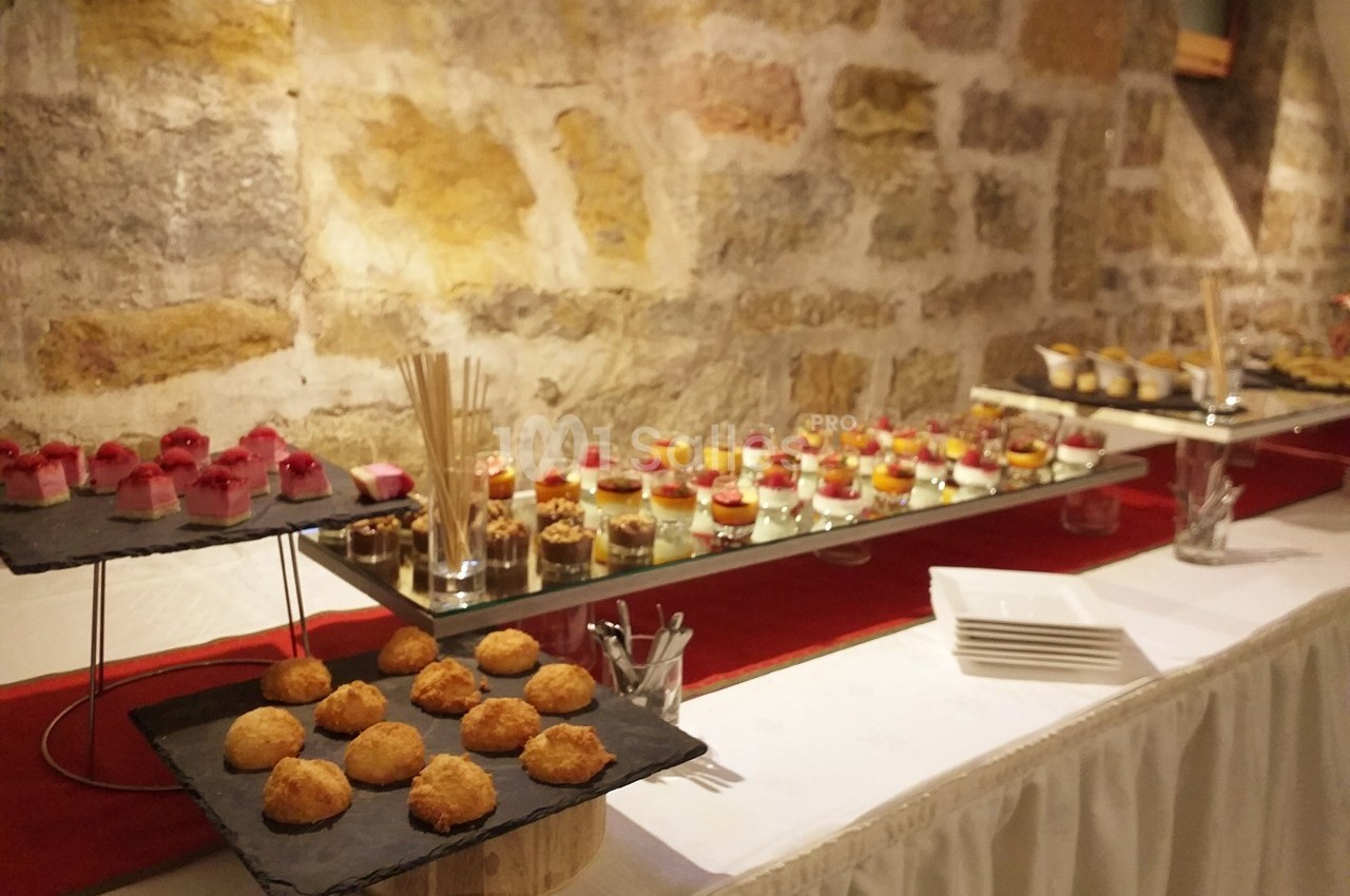 Buffet de desserts variés disposés sur des plateaux devant un mur en pierre, avec une nappe blanche.