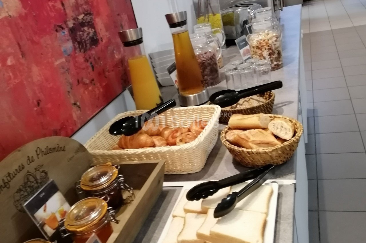 Buffet de petit-déjeuner avec viennoiseries, pain, confitures, jus de fruits et céréales disposés sur une table.