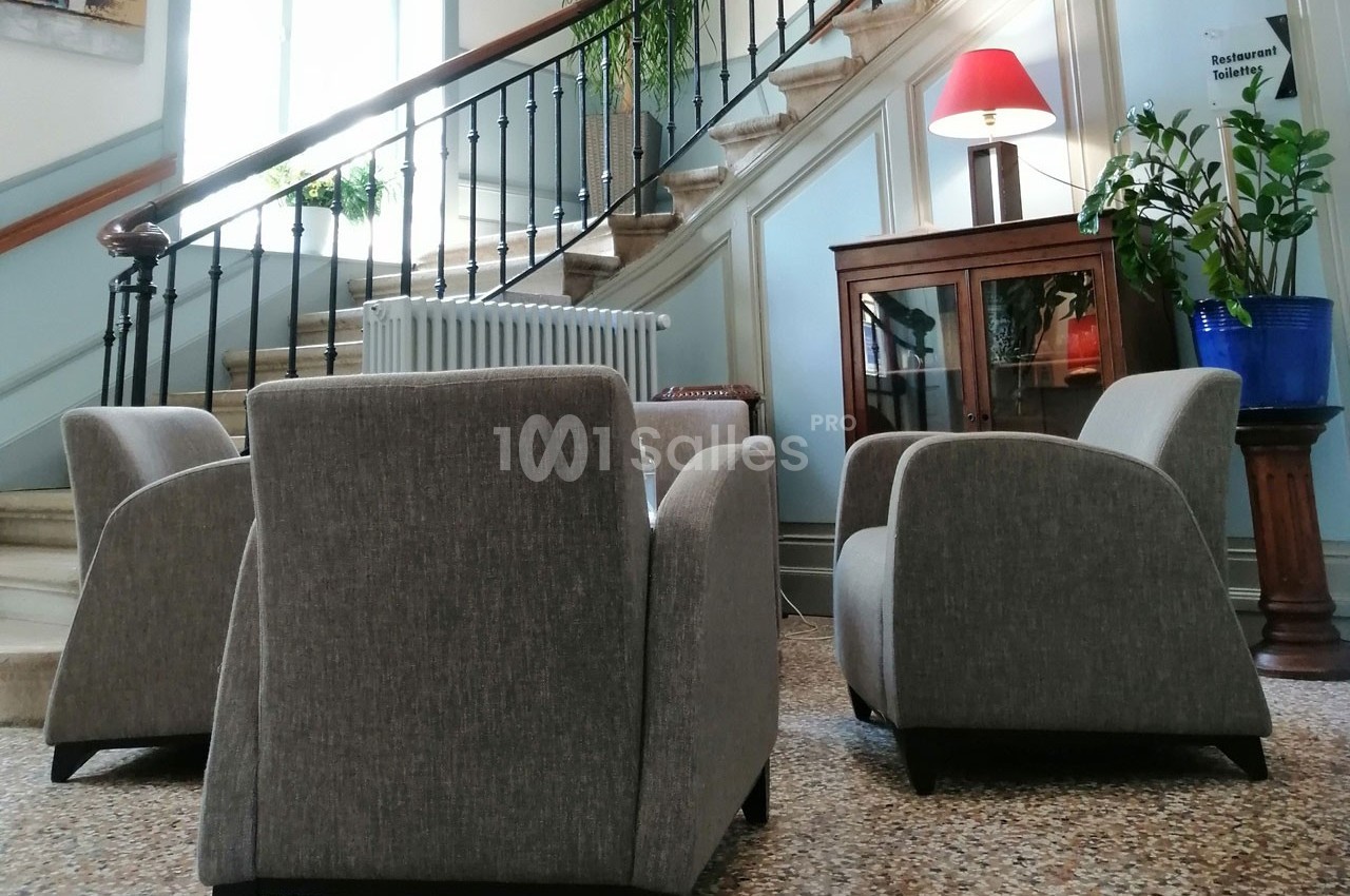 Trois fauteuils gris disposés autour d'une table basse dans un hall avec escalier en colimaçon et plantes décoratives.