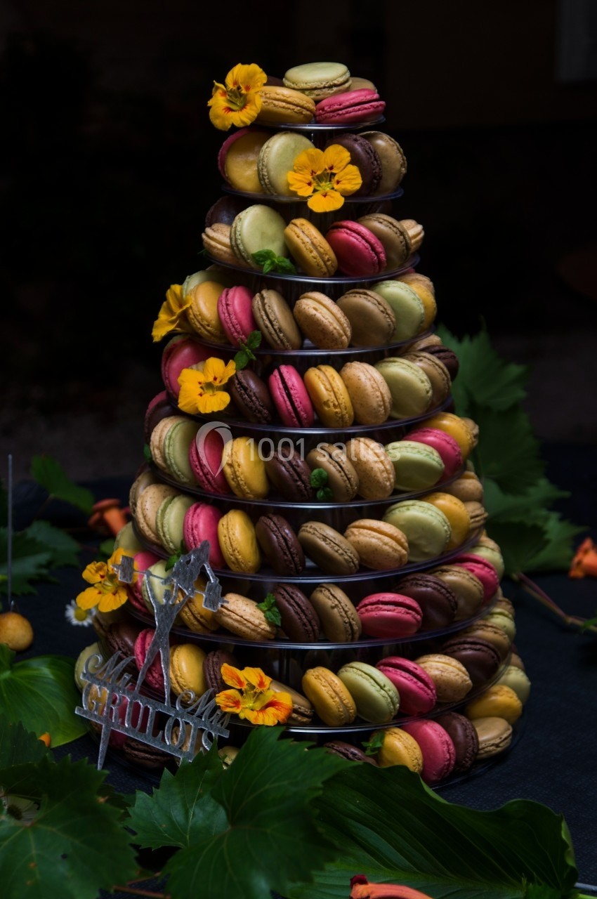 Tour de macarons colorés décorée de fleurs jaunes, posée sur une table sombre avec des feuilles en arrière-plan.