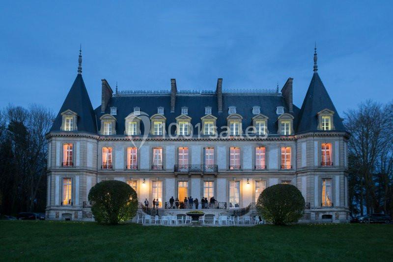 Façade éclairée d'un château de style classique au crépuscule, avec des invités visibles sur la terrasse.