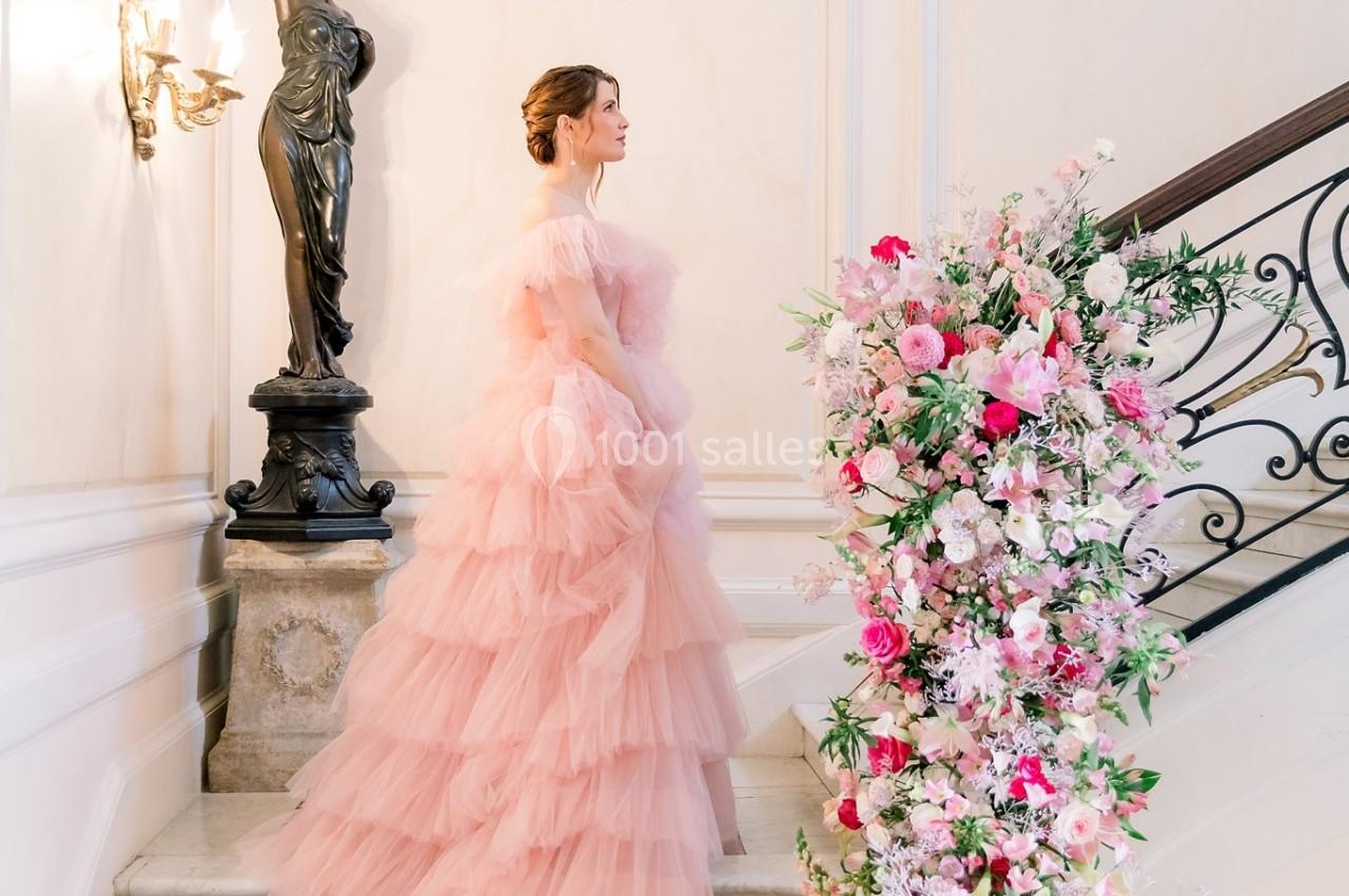 Femme en robe rose volumineuse montant un escalier décoré de fleurs colorées, dans un intérieur élégant.