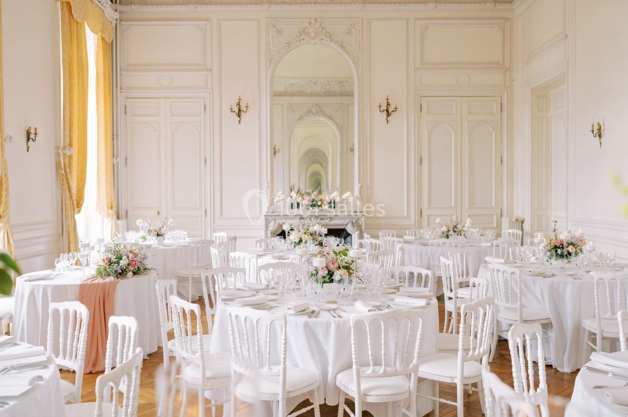 Salle de réception élégante avec tables rondes dressées, nappes blanches et décorations florales pastel.