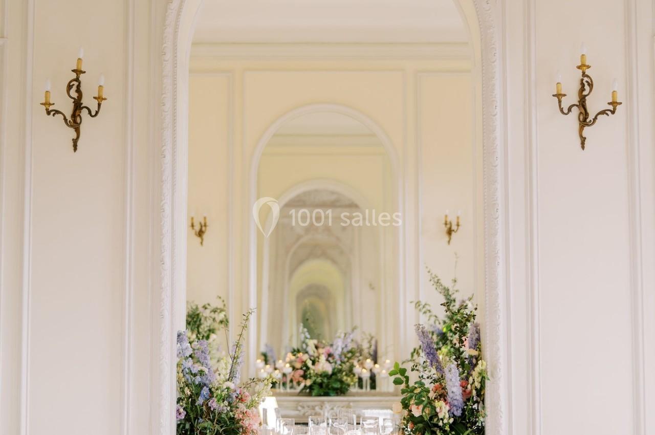 Salle élégante avec moulures, miroir central reflétant des compositions florales, tables dressées et chaises blanches.