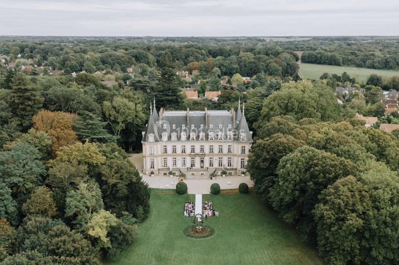 Vue aérienne d'un château entouré de jardins et d'arbres, avec une cérémonie en plein air sur la pelouse.