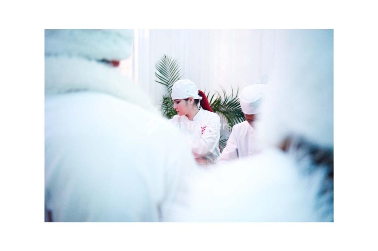 Une cheffe cuisinière en tenue blanche travaille dans une cuisine, entourée de plantes et d'autres personnes.