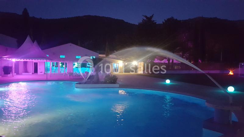 Piscine éclairée de nuit avec jets d'eau, entourée de bâtiments illuminés et de collines en arrière-plan.