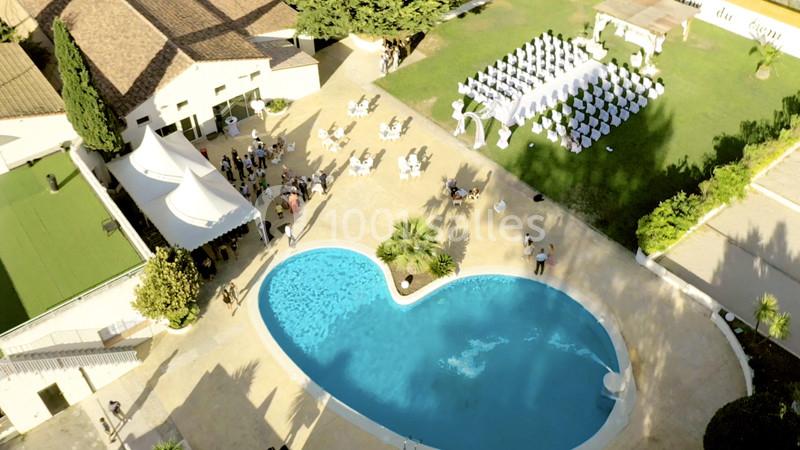 Vue aérienne d'un lieu de réception avec piscine en forme de cœur, espaces verts et chaises disposées pour une cérémonie.