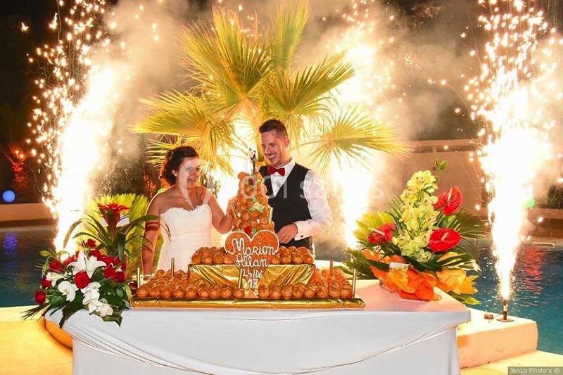 Un couple en tenue de mariage coupe une pièce montée devant une table décorée, avec des feux d'artifice en arrière-plan.