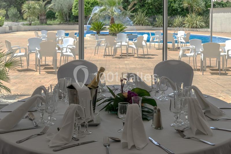 Table dressée avec nappes blanches, verres et couverts, vue sur une terrasse avec piscine et chaises en arrière-plan.