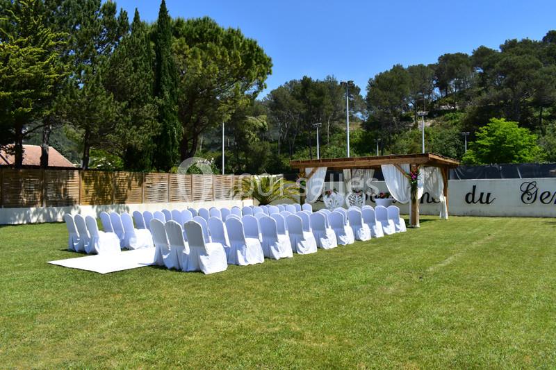 Chaises blanches alignées en extérieur devant une pergola décorée, sur une pelouse entourée d'arbres.