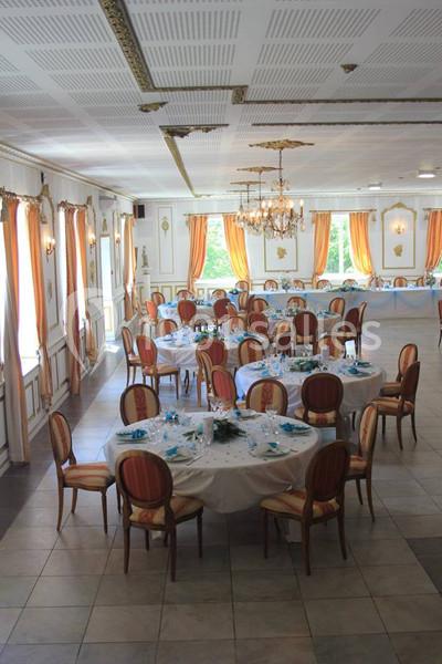Salle de réception élégante avec tables rondes dressées, chaises en bois, lustres et grandes fenêtres avec rideaux orange.
