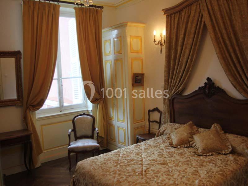 Chambre classique avec lit double, mobilier en bois, rideaux dorés et lumière naturelle entrant par une fenêtre.